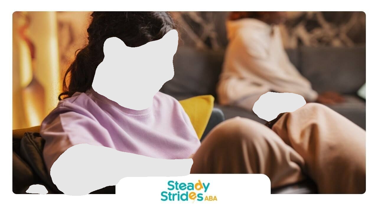 Woman in lilac shirt looks down, man blurred on couch. Interior.