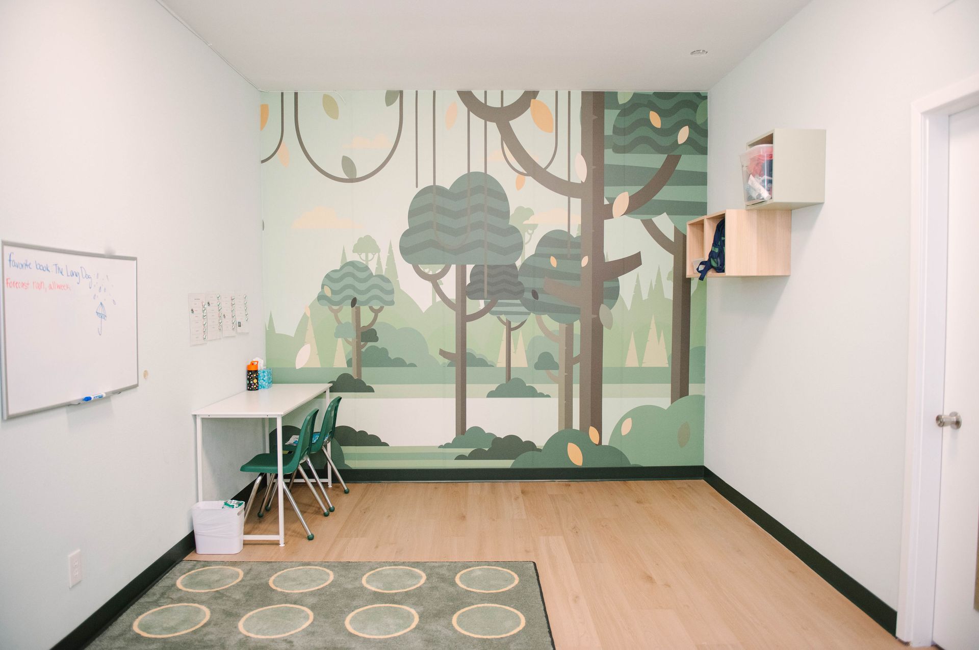 A room with a jungle mural, white walls, and wooden floor. There's a whiteboard, table, chairs, and rug.