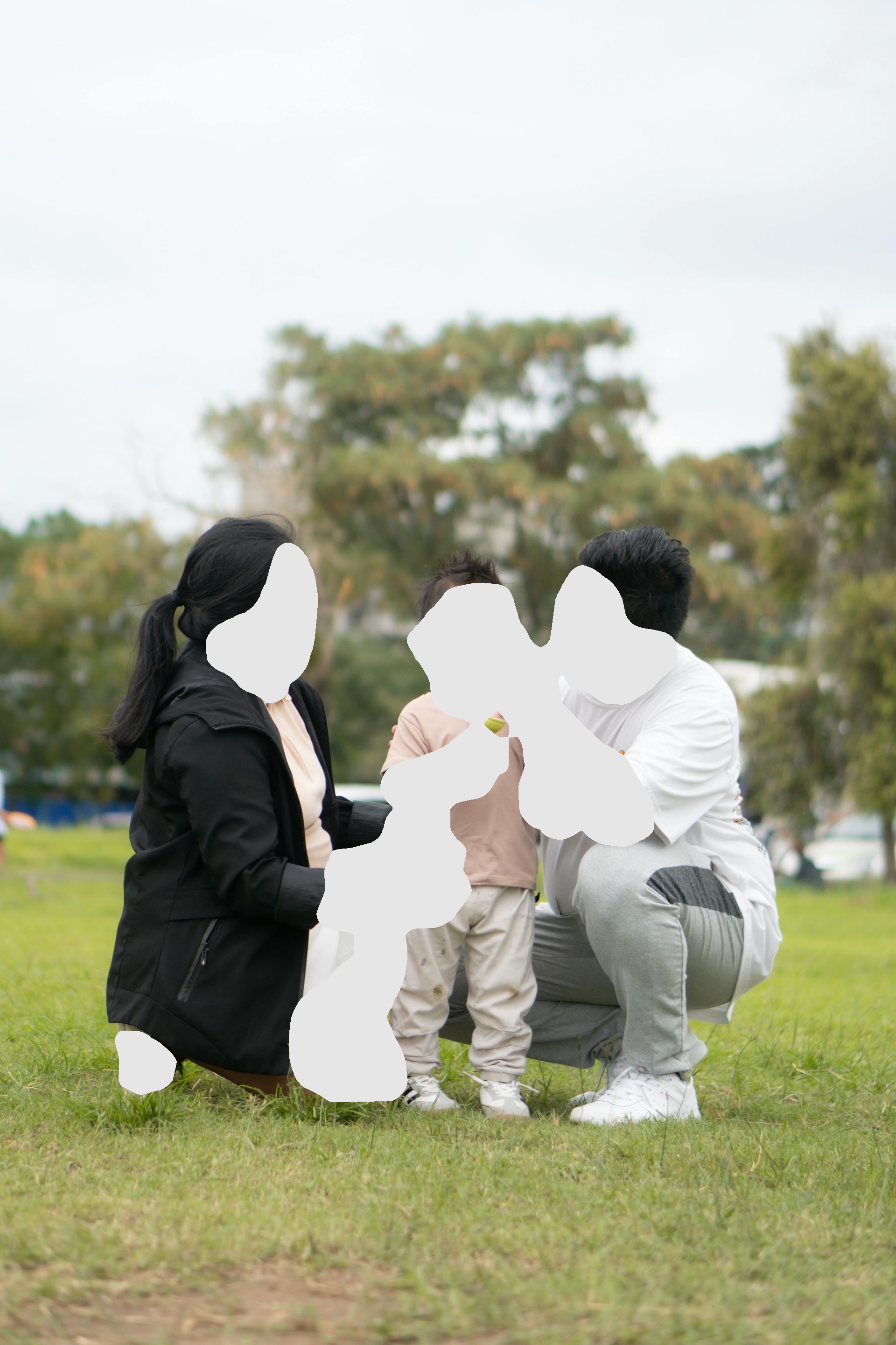 Family of three in park: father adjusts child's ear while mother smiles holding a snack.
