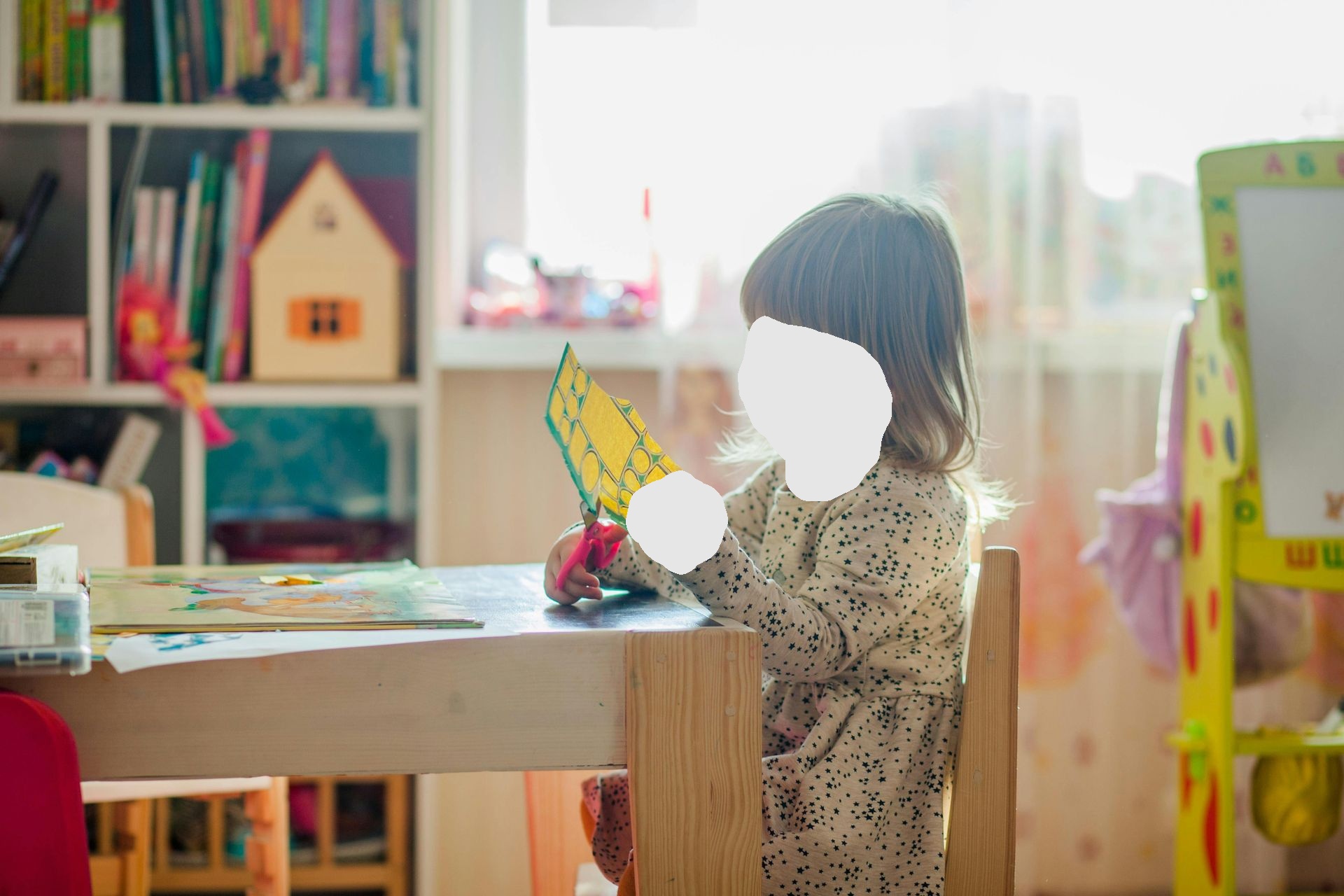 Young child seated at a table, cutting yellow paper with scissors in a well-lit room.